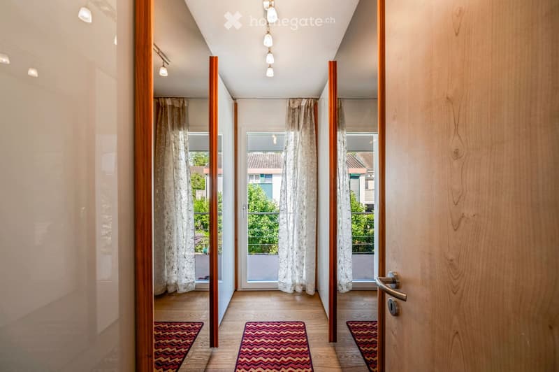 Bright hallway with wooden flooring, white walls, and three doors leading to rooms. Each door has a curtained window, and small patterned rugs are placed in front of each door.