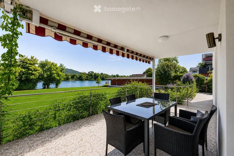 Balcony with a glass table and wicker chairs, overlooking a lake and green park area.