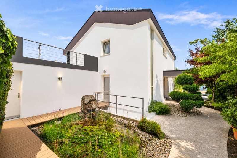 Modern two-story house with white exterior, black accents, and a small garden area featuring a pond, wooden deck, and greenery.