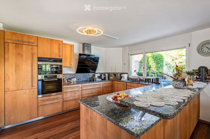 Modern kitchen with wooden cabinets, granite countertop, built-in oven, and stainless steel appliances. Features a central island with a bowl of fruit and decorative doilies.
