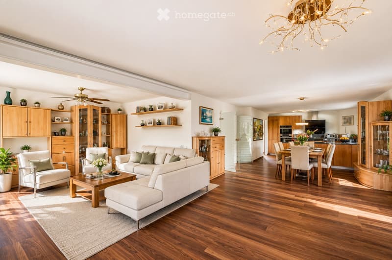 Open-plan living and dining area with light-colored furniture, wooden flooring, and modern decor including a chandelier and wall shelves.