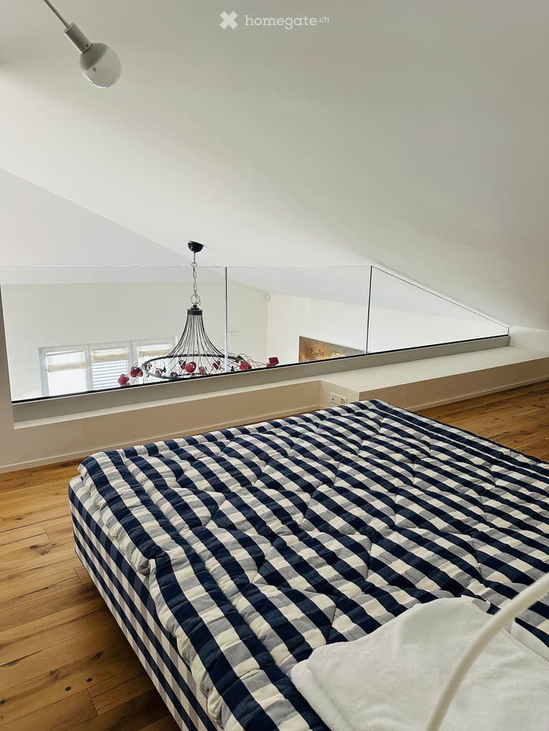 A modern bedroom with a large bed covered in a blue and white checkered quilt, wooden flooring, a chandelier hanging from the ceiling, and a large mirror reflecting part of the room.