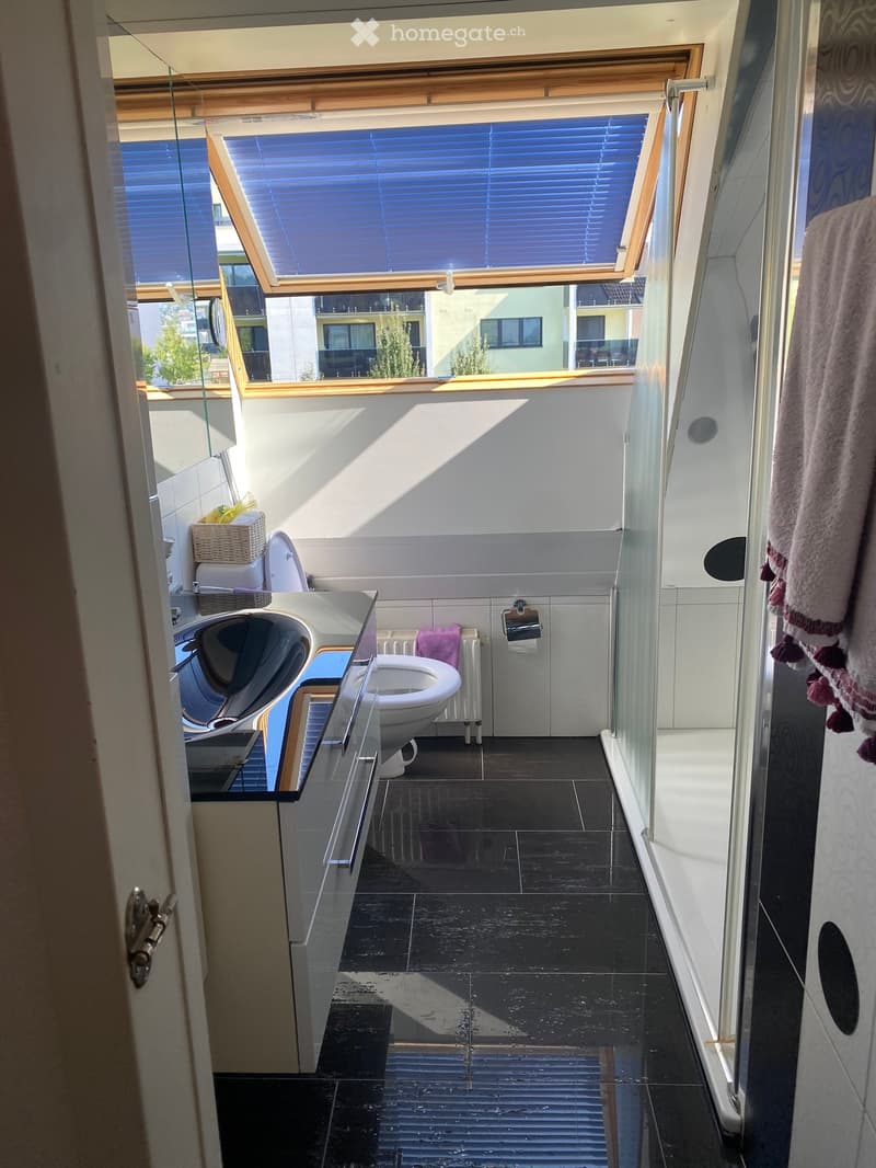 Modern bathroom with dark tiled floor, white walls, toilet, sink, radiator, and a towel hanging on the side. A large window with blinds offers a view of the outside.