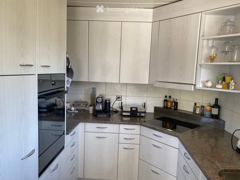 Modern kitchen with white cabinets, granite countertops, and various appliances including a refrigerator, oven, coffee maker, and toaster. Shelves hold decorative items and bottles.