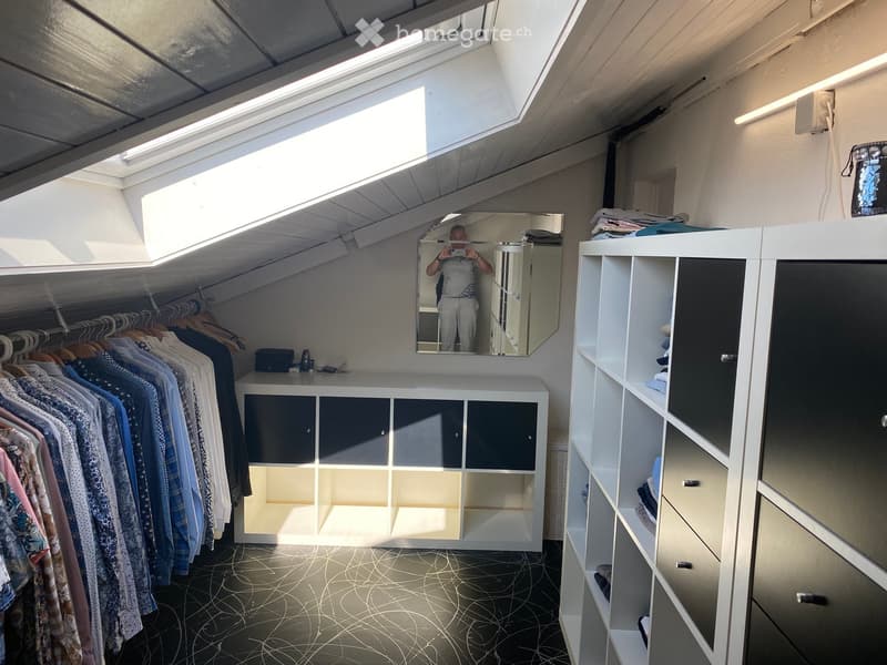 Well-lit walk-in closet with white built-in wardrobe, clothing rack, and large mirror. Features modern design with dark flooring and ample storage space.
