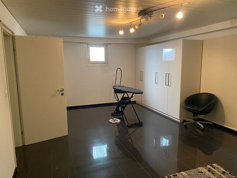A small, well-lit room with dark tiled flooring, white walls, and built-in white cabinets. Contains a black swivel chair, an ironing board, and a small rug near the door.