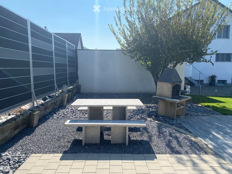Well-maintained garden with a stone table, benches, and a barbecue, surrounded by gravel and a modern fence.