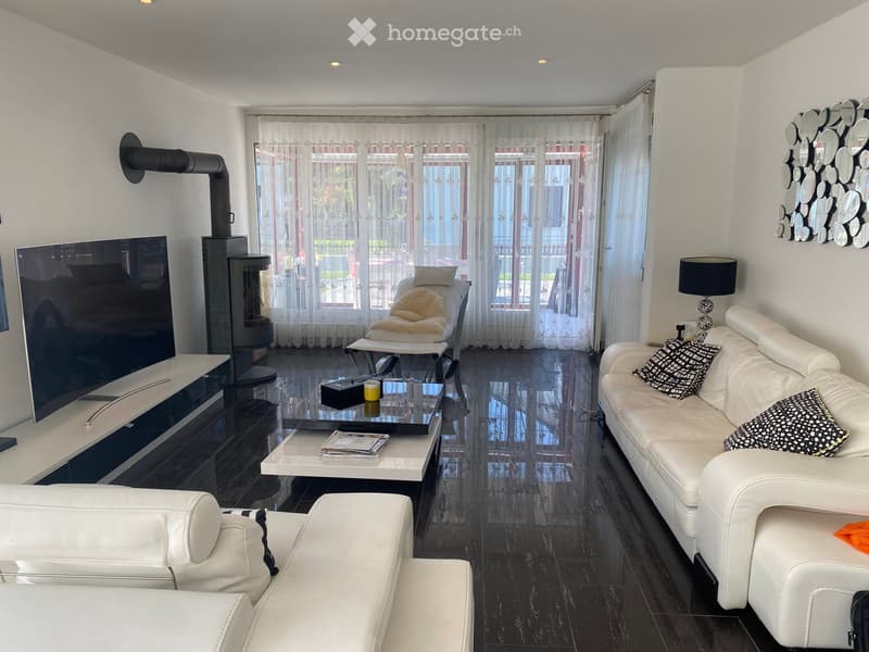 Modern living room with white leather sofa, coffee table, fireplace, large windows with sheer curtains, and a television on a stand.