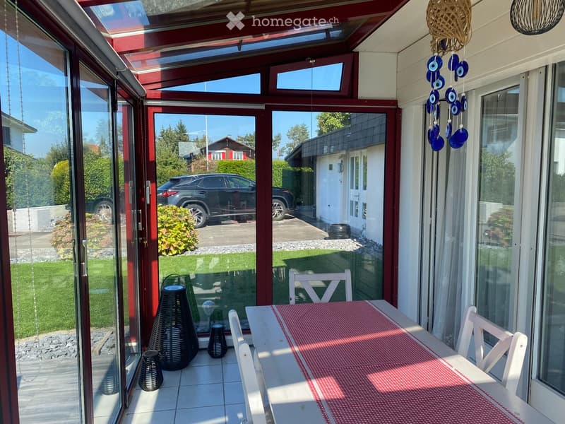 Bright, enclosed patio with white table, red tablecloth, white chairs, and decorative wicker lamps. Large glass doors open to a well-maintained garden and driveway with a parked car.