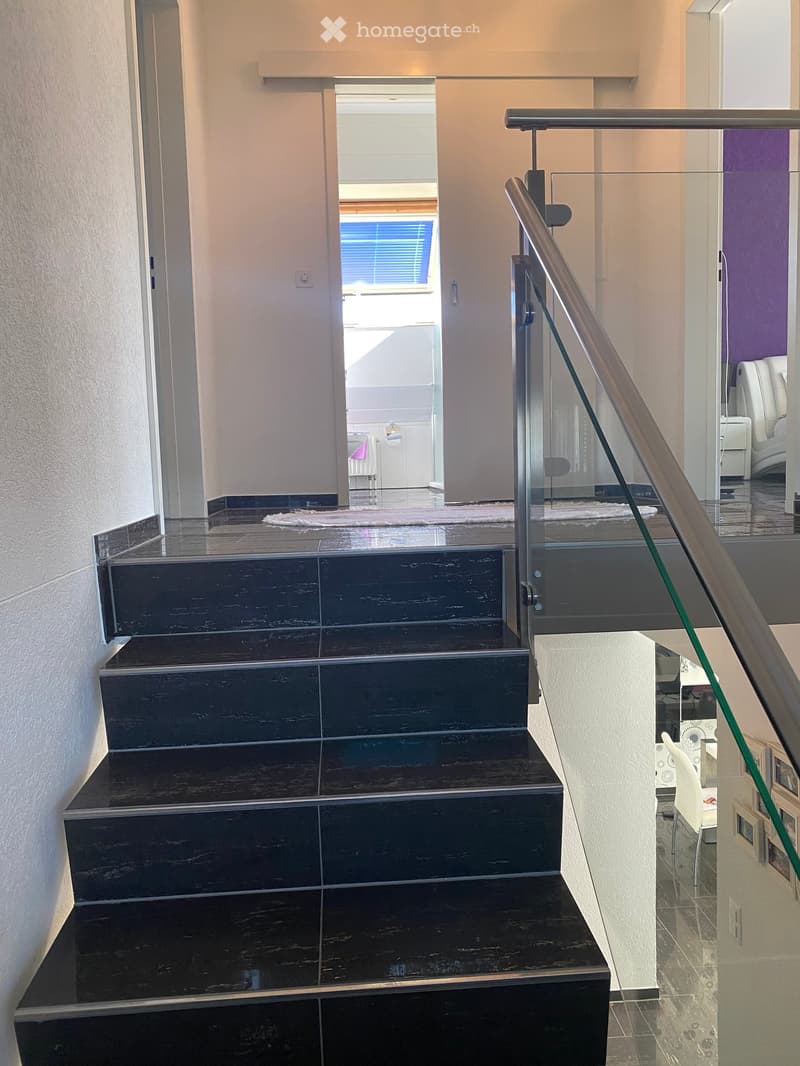 Modern staircase with dark wooden steps and a glass railing, leading to a bright room with white furniture and a purple accent wall.