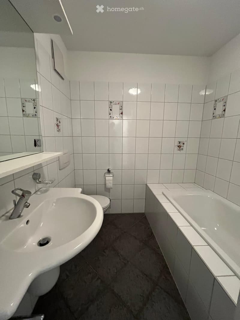 A small bathroom with white tiled walls, a white sink with a chrome faucet, a toilet, a bathtub, and a mirror above the sink.
