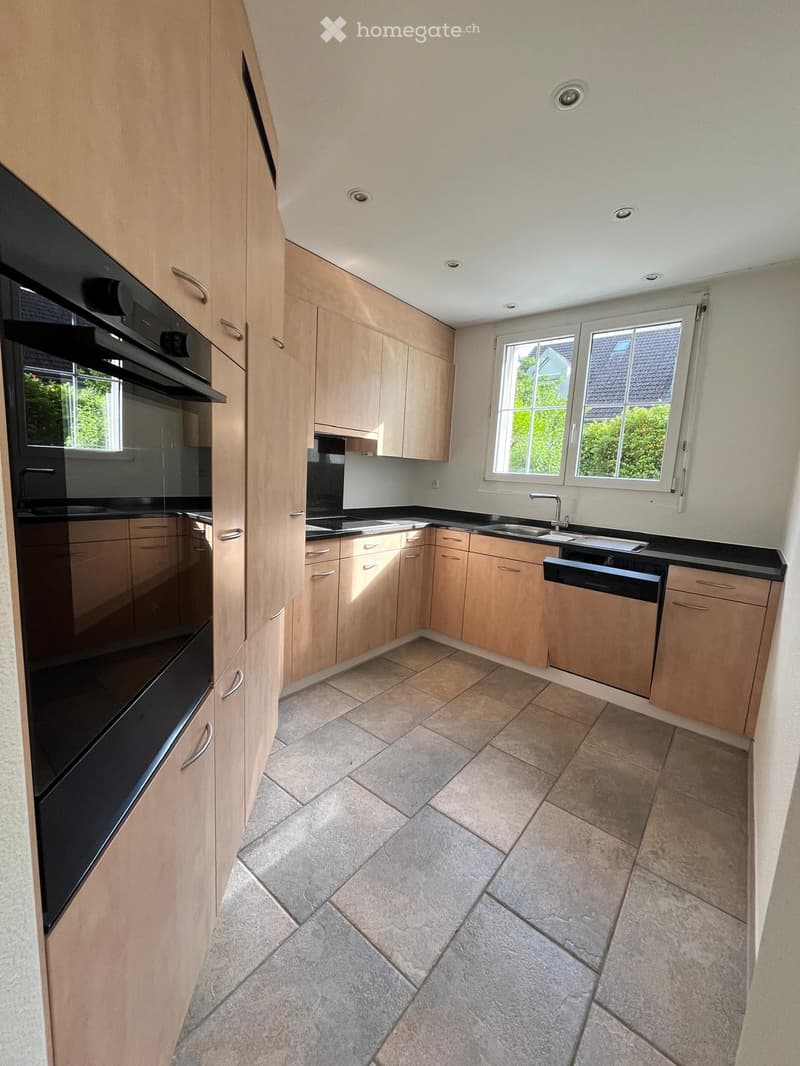 Modern kitchen with light wooden cabinets, black countertops, tiled floor, and stainless steel appliances including an oven, sink, and dishwasher.
