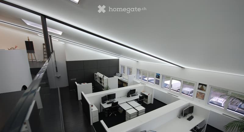 Modern office space with white desks, black chairs, multiple computer setups, and large windows overlooking the outside.
