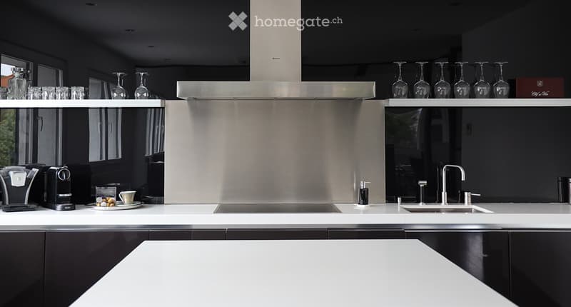 Modern kitchen with stainless steel appliances, white countertops, and black cabinets. Features a coffee machine, wine glasses, and a sink with a modern faucet.