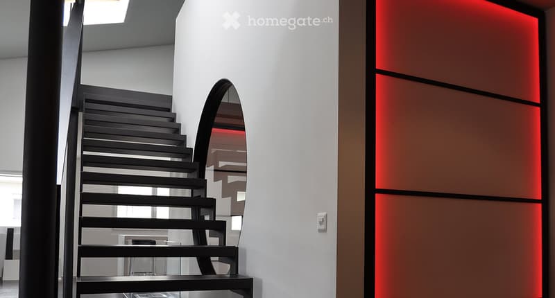 Modern hallway with a sleek black staircase, illuminated red wall panel, and a large mirror reflecting the staircase.