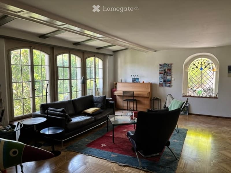 Bright living room with large windows, dark wooden floor, black leather sofa, coffee table, chairs, and a piano.