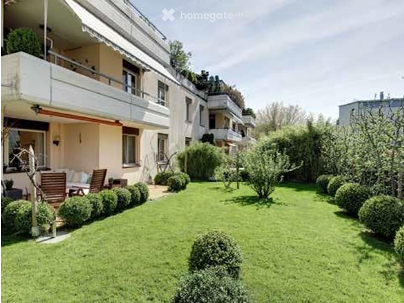 Well-maintained garden area with neatly trimmed hedges, a small seating area with chairs and a table, and a multi-story building with balconies in the background.