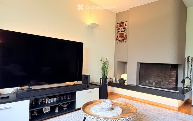 Modern living room with a large flat-screen TV, a sleek black entertainment unit, a round wooden coffee table with a decorative centerpiece, a built-in fireplace with a brick surround, and a wall-mounted shelf with decorative items.
