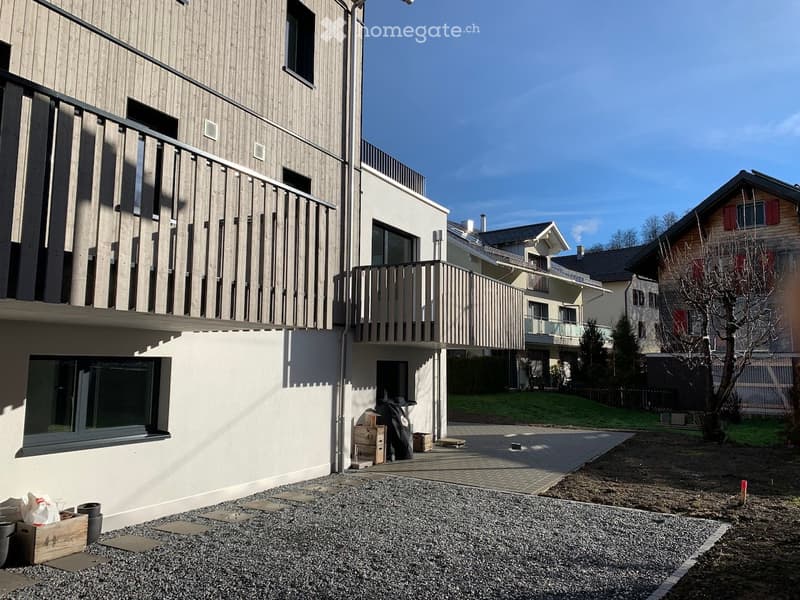 Modern multi-story residential building with balconies, large windows, and a paved driveway. The building features a combination of wood and white exterior finishes.