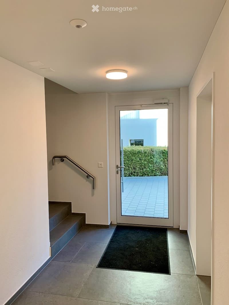 Modern hallway with tiled floor, white walls, recessed lighting, a staircase with a metal handrail, and a glass door leading to an outdoor area.