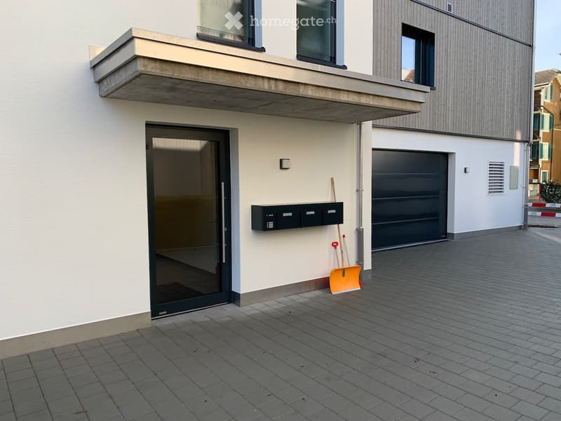 Modern building entrance with a large glass door, a black garage door, and a small balcony above. A snow shovel is leaning against the wall near the entrance.