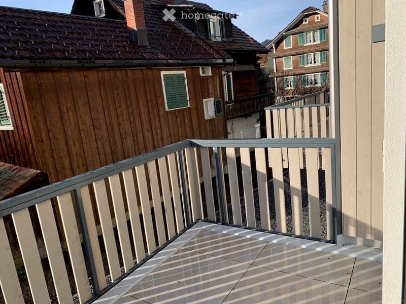 Balcony with metal railing, tiled floor, overlooking a residential area with traditional houses and wooden facades.