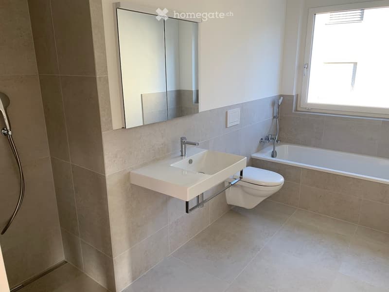 Modern bathroom with a white sink, toilet, bathtub, and large mirror above the sink. Light-colored tiles and a window provide natural light.