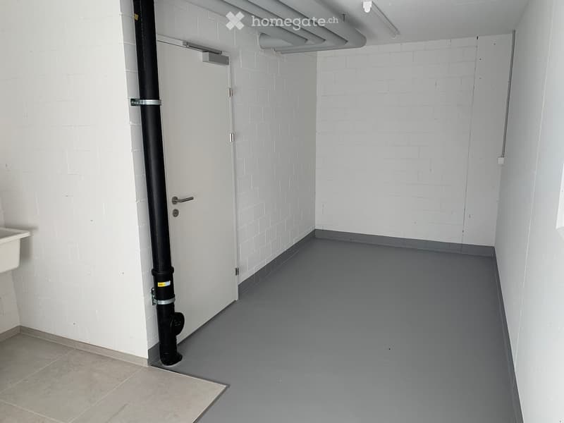 Empty room with white brick walls, grey floor, and a large black pipe running vertically along one wall. A white door is visible at the far end.