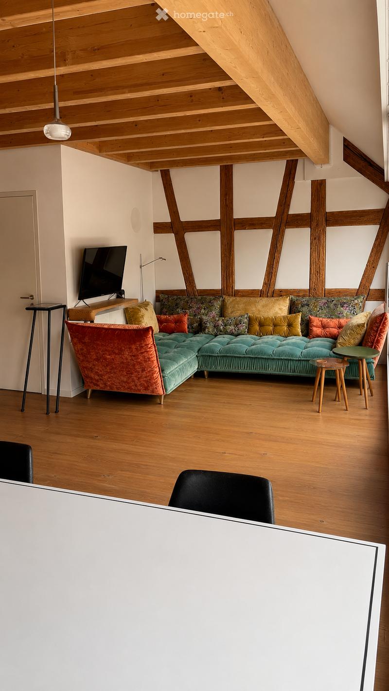 Modern living room with wooden floors, exposed wooden beams, a sectional sofa with colorful cushions, a small table, and a television on a stand.