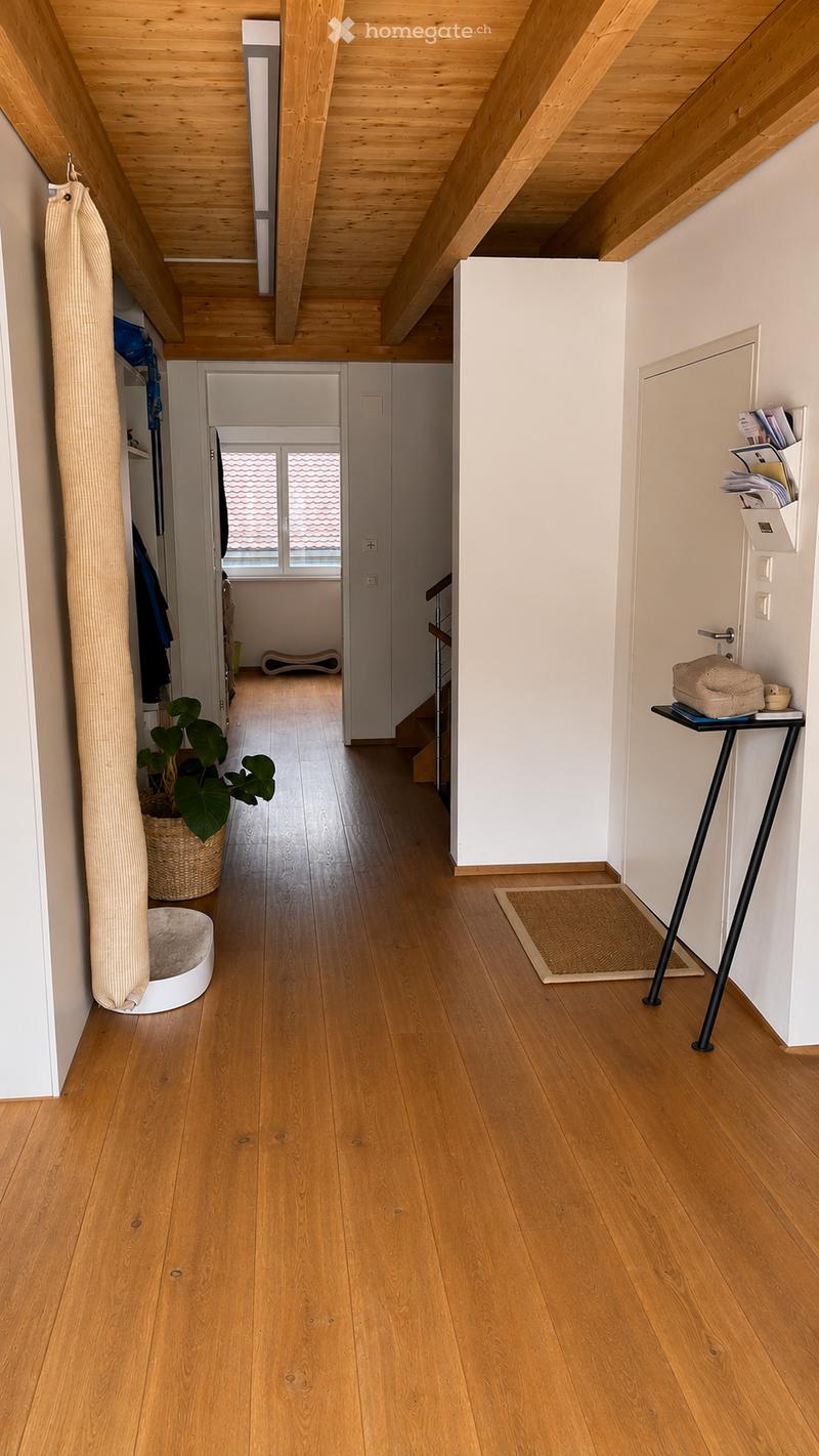 Bright hallway with wooden flooring, white walls, and exposed wooden beams on the ceiling. Features a small table with a plant, a pet bed, and a coat rack with hanging items.