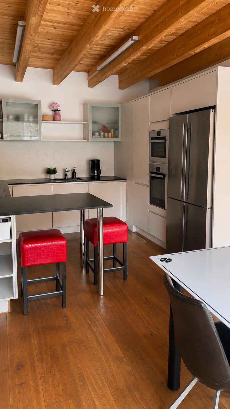 Modern kitchen with light-colored cabinets, stainless steel appliances, wooden flooring, and a black countertop. Features include a coffee maker, sink, two red stools, and a small dining area with a table and chair.