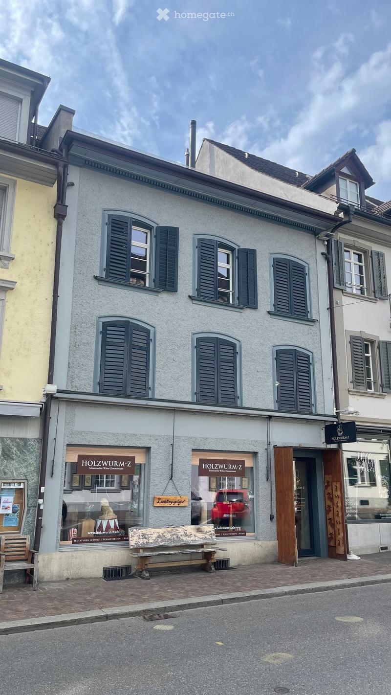 Two-story building with light grey facade, multiple windows with dark shutters, and a storefront labeled "HOLZWURM-Z" with a bench and a small wooden chair outside.