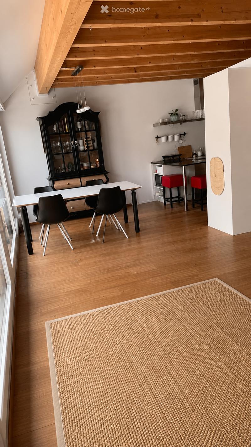 A modern, open-concept room with a dining area and a small kitchen. The space features a white dining table with four black chairs, a black glass cabinet, a small kitchen counter with a microwave, red stools, and pendant lighting.