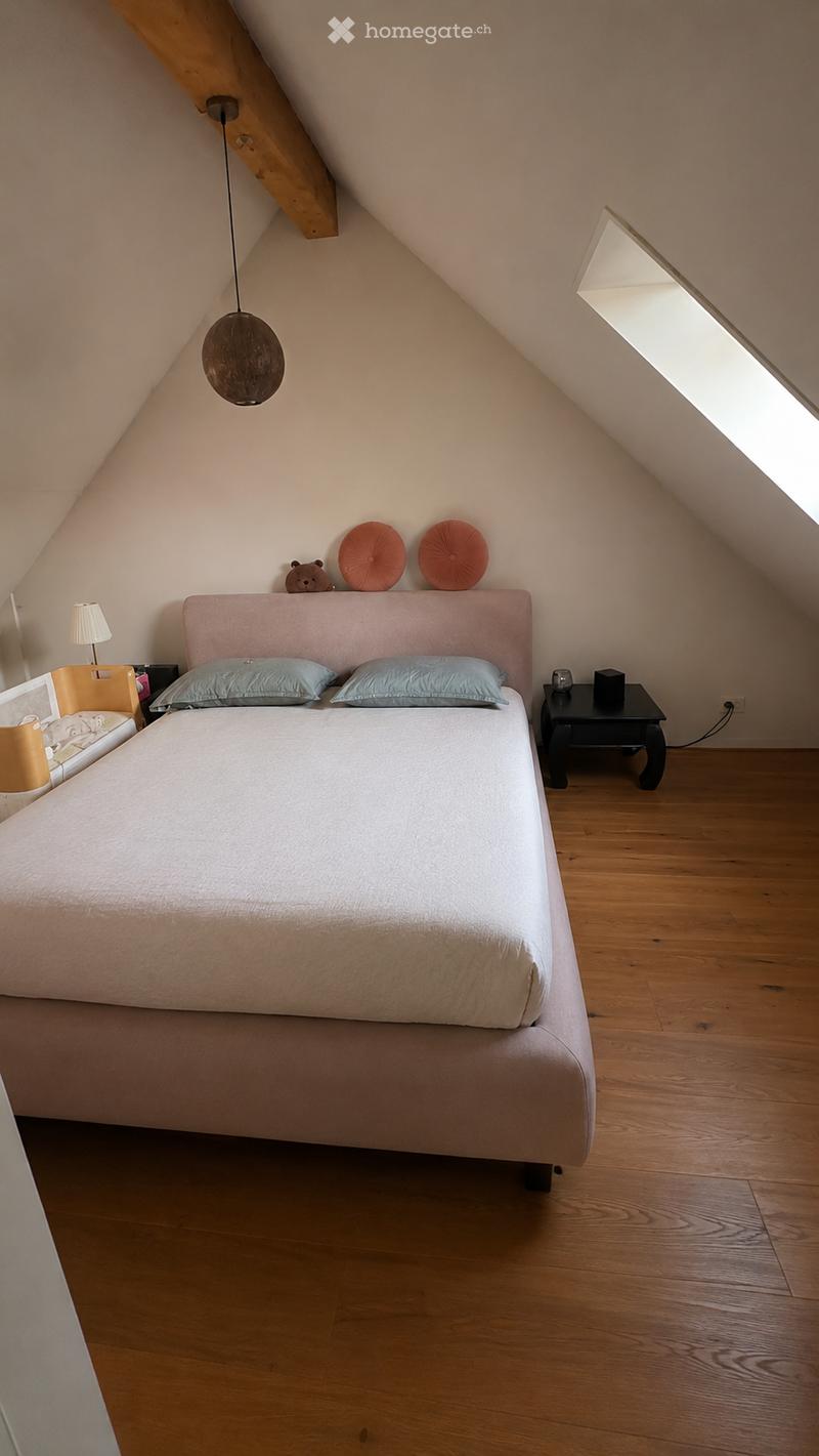 Cozy attic bedroom with wooden flooring, slanted ceilings, and a large bed with light-colored bedding. Features a nightstand with a table lamp, a pendant light, and decorative pillows on the headboard.