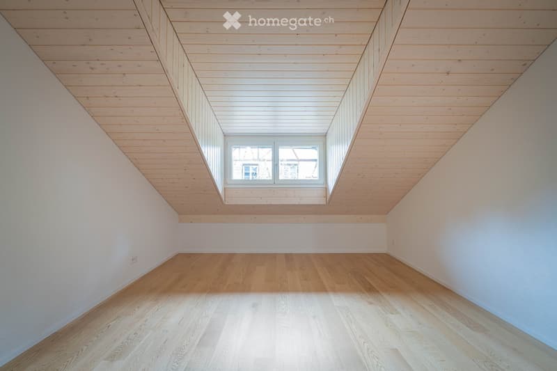 Empty attic room with wooden floors and slanted wooden ceilings, featuring a single window with white frame and glass panes.