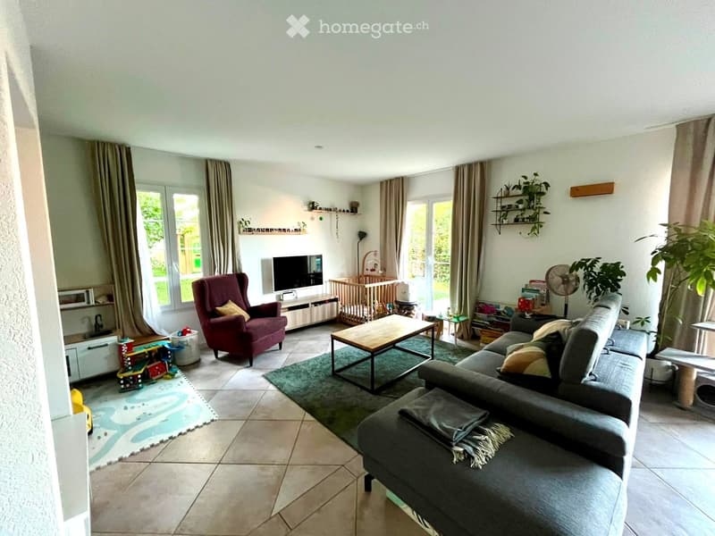 Bright living room with large windows, tiled floor, grey sectional sofa, wooden coffee table, red armchair, TV, baby crib, and decorative shelves.