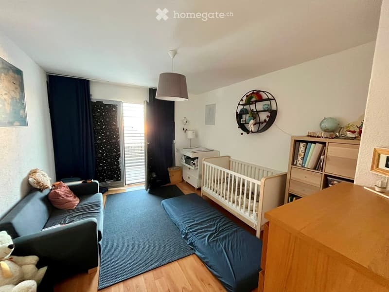 A cozy, well-furnished room with a blue sofa, a crib, a bookshelf, a small table, and a hanging lamp. The room features a large window with dark curtains and a view of the outside.