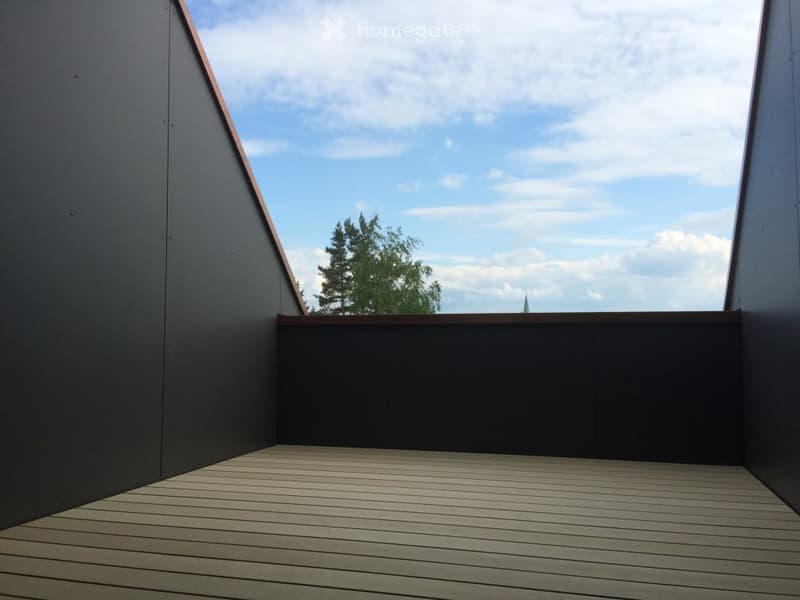 Modern rooftop terrace with wooden decking, dark grey walls, and a view of trees and sky.