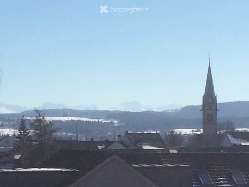 A view of a town with traditional houses and a prominent church tower in the background, surrounded by snow-covered hills and a clear blue sky.