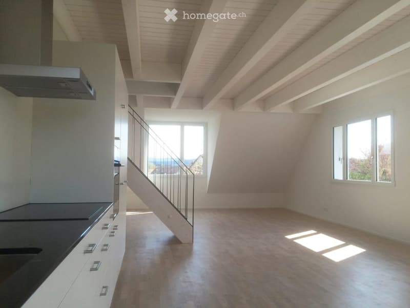 Open-plan space with a modern kitchen area and a staircase leading to an upper level. The room features white walls, wooden flooring, and large windows allowing natural light to fill the space.