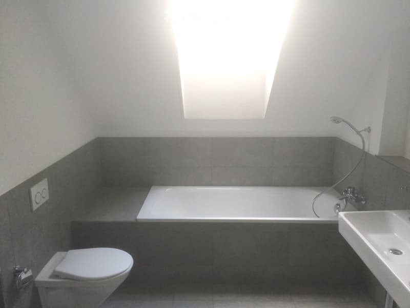 Modern bathroom with a bathtub, toilet, sink, and a window above the tub. The walls are tiled in a dark gray color, and the floor is tiled in a light gray color.