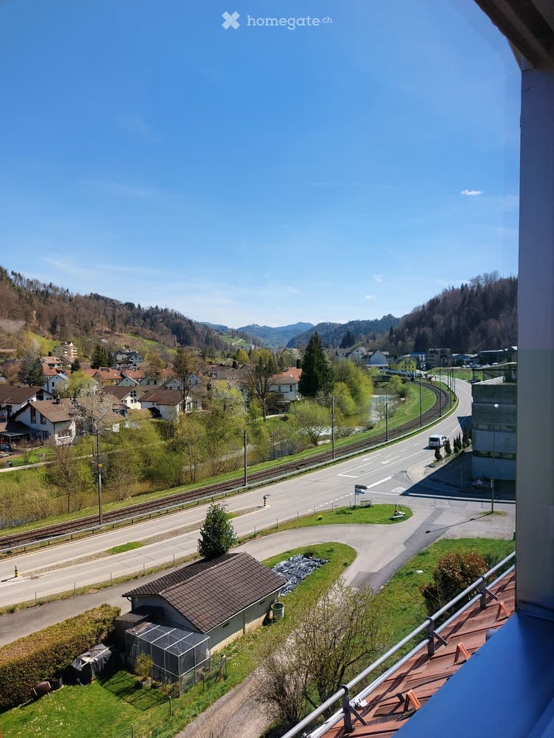 View from a balcony showing a town with houses, roads, a railway line, and surrounding hills under a clear blue sky.