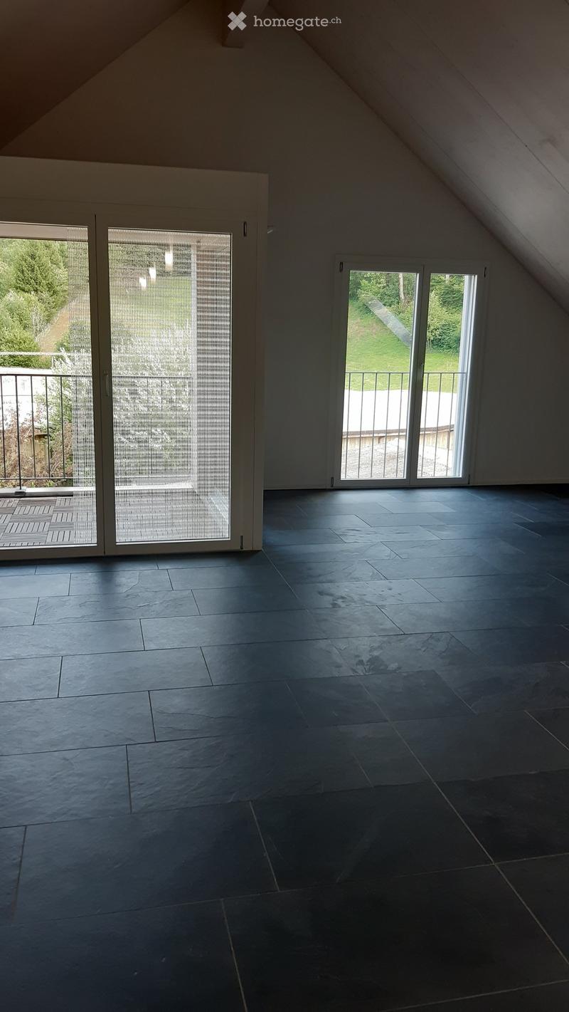 An empty attic room with dark tiled flooring, large windows, and sliding glass doors leading to a balcony.