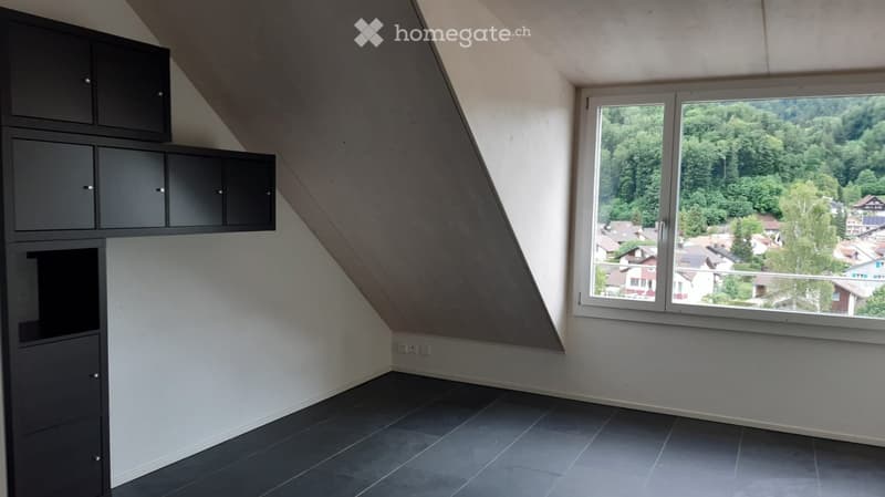 An empty attic room with slanted ceilings, dark grey tiled floor, built-in black cabinets, and large windows offering a scenic view of a town and greenery.