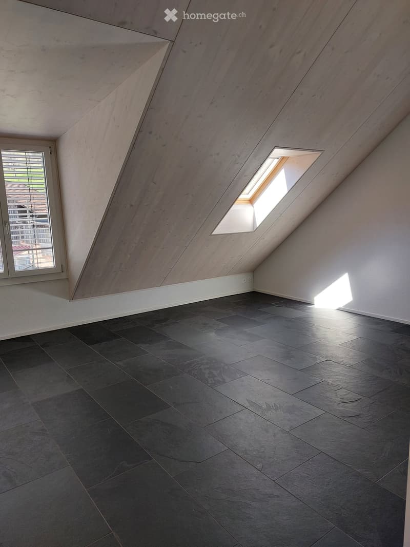 An empty attic room with dark grey tiled flooring, white walls, and a slanted wooden ceiling. Features include a large skylight window and a smaller window with blinds on the left side.