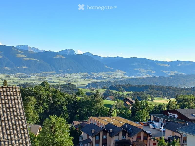 Panoramic view of a village with houses, trees, and distant mountains under a clear blue sky.