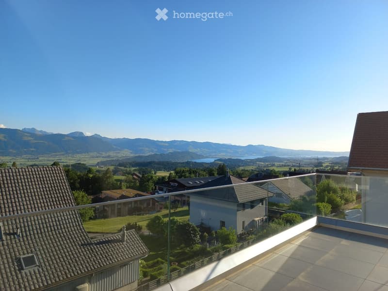 Balcony with glass railing, overlooking a scenic landscape with mountains, fields, and houses.