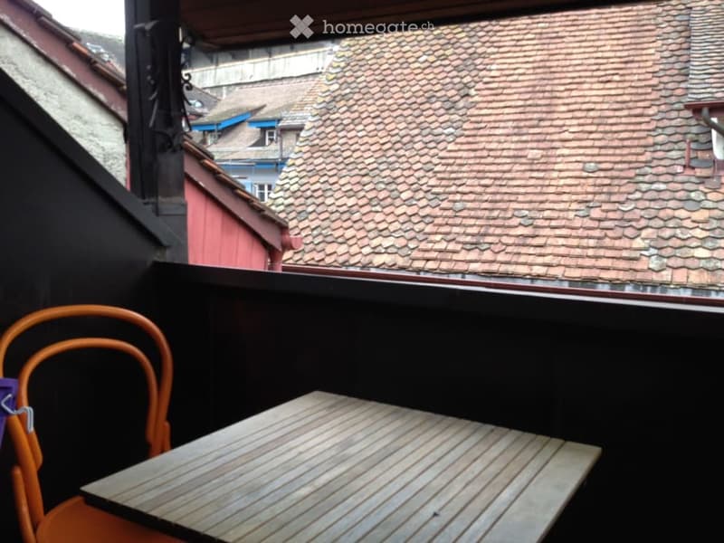 Balcony with wooden table and orange chair, overlooking a tiled roof area with neighboring buildings.