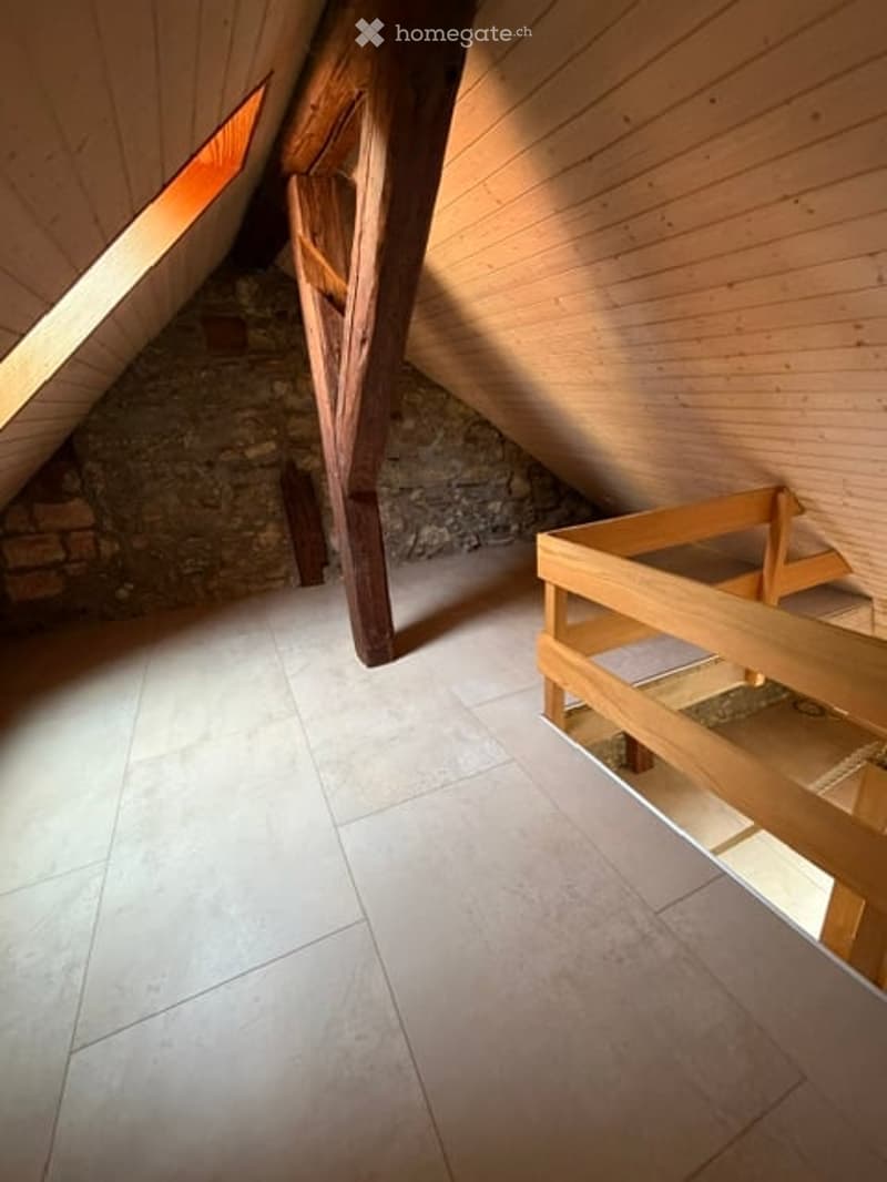 An attic space with wooden beams, exposed brick wall, and a small window letting in natural light. The floor is tiled, and there is a wooden platform or loft area on the right side.