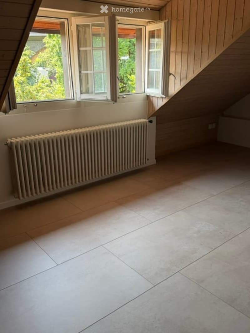 Empty attic room with wooden slanted ceilings, large windows, tiled floor, and radiator.